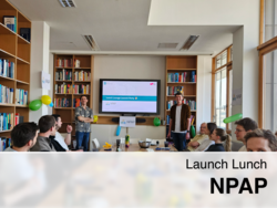 Ein Team sitzt in einem hellen Bibliotheksraum mit bunten Luftballons, während zwei Personen vor einem Bildschirm mit der Aufschrift „Lunch Lounge Launch Party NPAP“ präsentieren.