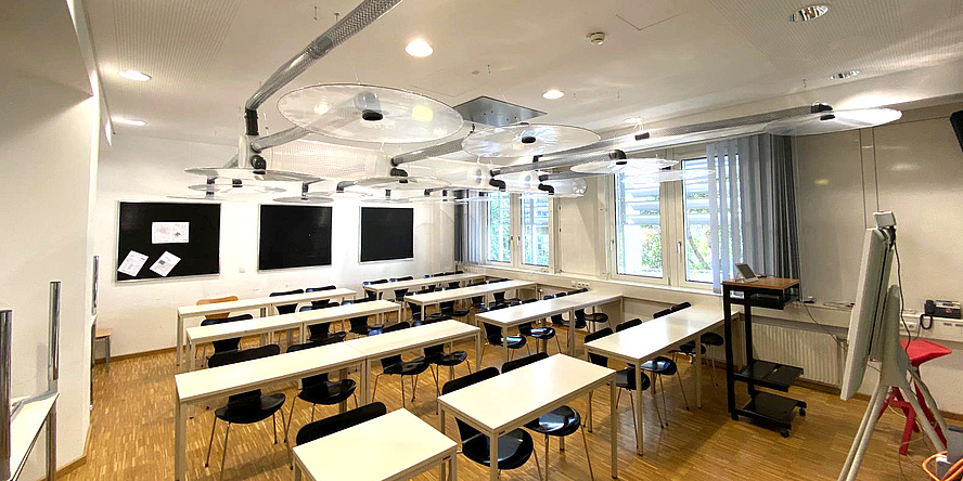 View into a classroom, under the ceiling pipes and circular transparent plate-like objects can be seen.