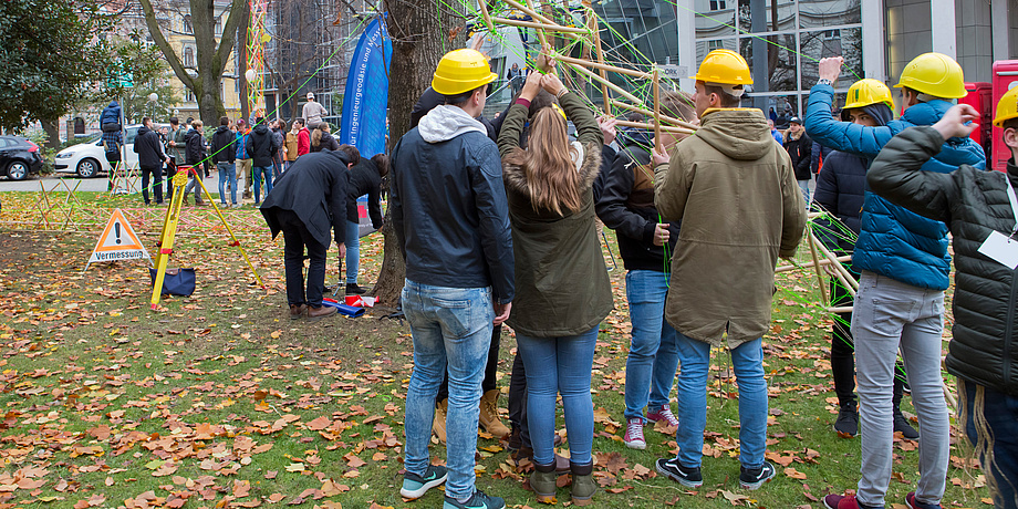 Schülerinnen und Schüler bauen auf einer belaubten Wiese hinter der alten Technik gemeinsam an einem Turm aus grünem Stabwerk.