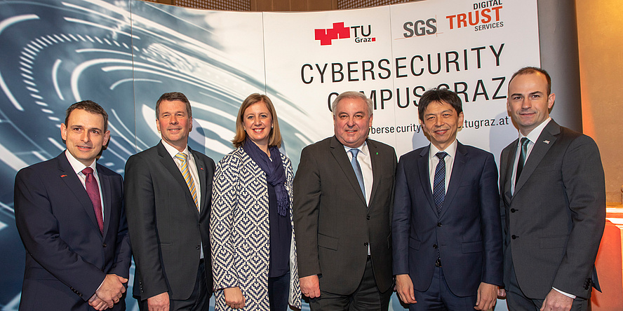 Five men and a woman are standing in front of an expo wall, on which the logos of TU Graz, SGS and the URL of the Cybersecurity Campus Graz can be read.