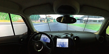 Ein Auto von innen. Blick von der hinteren Sitzbank aus. Vor dem Fahrenden ist ein Lenkrad sichtbar. Vorne ist die Windschutzscheibe, vor der Bildschirme angebracht sind. Auf den Bildschirmen wird eine Straße simuliert.