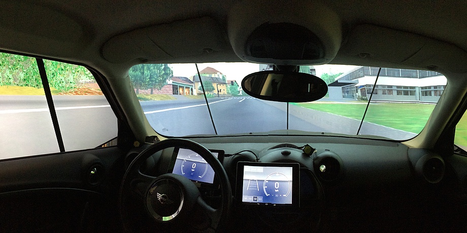 Ein Auto von innen. Blick von der hinteren Sitzbank aus. Vor dem Fahrenden ist ein Lenkrad sichtbar. Vorne ist die Windschutzscheibe, vor der Bildschirme angebracht sind. Auf den Bildschirmen wird eine Straße simuliert.
