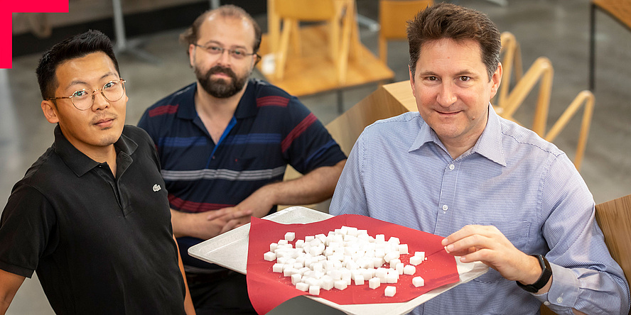 Three men look into the camera. One is holding a tray with lots of sugar cubes