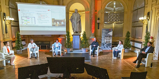 Sechs Personen sitzen in einem Halbkreis auf Stühlen in einem großen Raum. Vor ihnen stehen Monitore, dahinter steht eine Leinwand mit einer Präsentation.