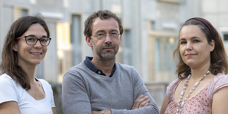 Drei Personen blicken in die Kamera. Links eine Frau mit braunen Haaren, weißem T-Shirt und einer Brille. In der MItte ein Mann mit braunen Haaren, einem grauen Pullover und einer Brille. Rechts eine Frau mit braunen Haaren, einem Haarband und einer bunten Bluse.