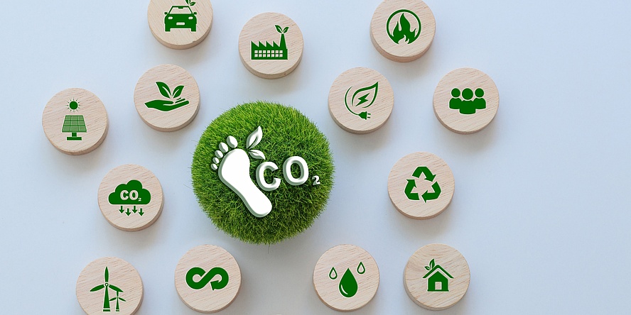 The image shows a central CO₂ footprint symbol, surrounded by 14 wooden discs featuring green icons related to environmental topics such as renewable energy, recycling, water, mobility, and sustainable practices.