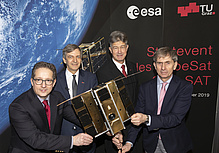 Vier MÄnner in dunklen Anzügen blicken in die Kamera und halten einen Satelliten in den Händen. 