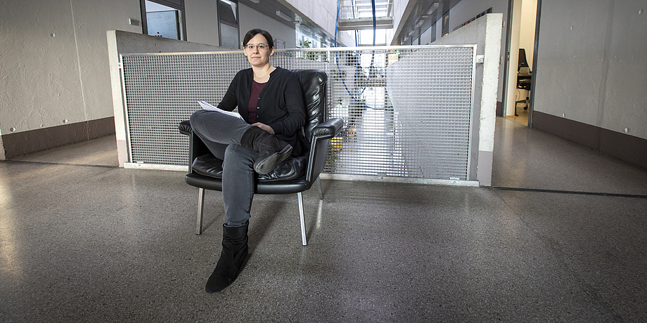 A young woman is sitting on a black leather armchair in front of a corridor. She has crossed her legs and looks directly into the camera. She has several sheets of paper in her hand.