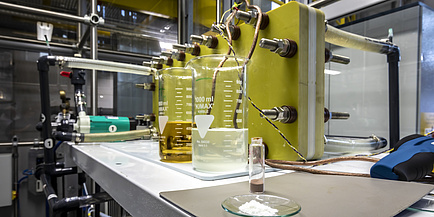 A small bowl with a powder, a small glass with a powder and two large glasses with a liquid stand in front of an experimental setup on a table