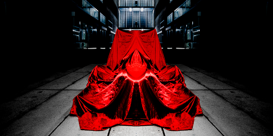 In einer Fabrikshalle steht im Zentrum ein Rennauto, dass von einem roten Samttuch verhüllt wird, sodass man nur die Silhouette sieht.