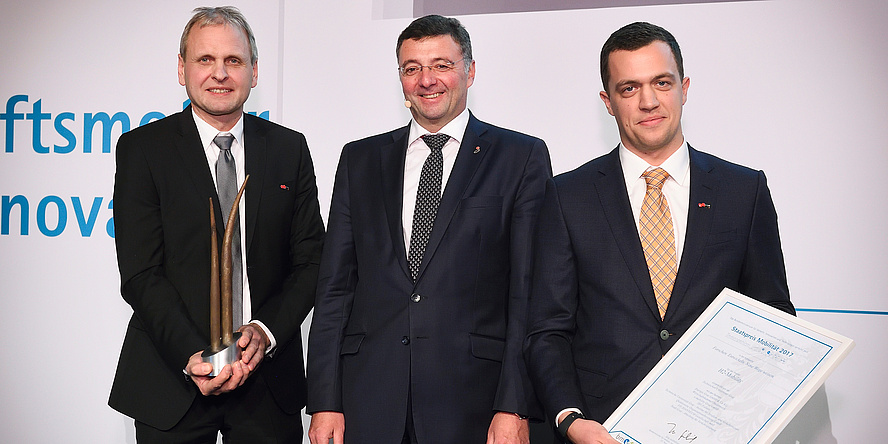 Three men in black suits smiling at the camera. One of them is holding a white piece of paper in his hands.