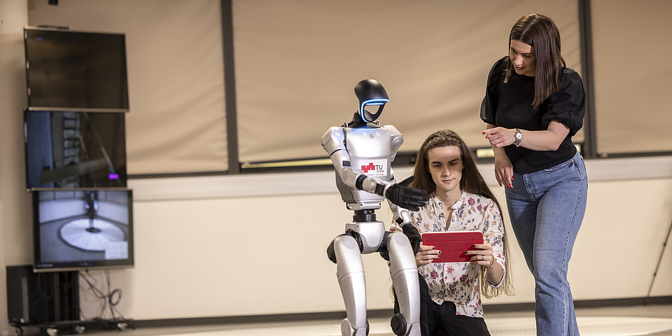 Zwei junge Menschen und ein humanoider Roboter. Eine der Personen hockt rechts neben dem Roboter, die andere Person steht rechts daneben. Die hockende Person hält ein Tablet, der Roboter und die stehende Person zeigen mit den Fingern darauf.