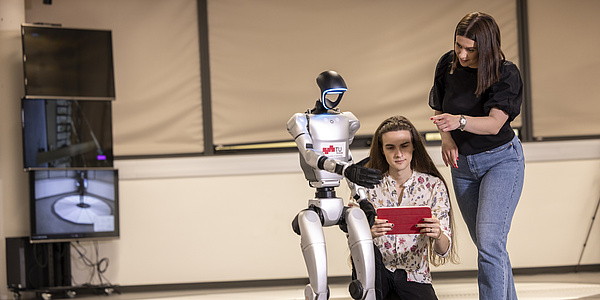 Two young people and a humanoid robot. One of the people is squatting to the right of the robot, the other person is standing to the right. The crouching person is holding a tablet, the robot and the standing person are pointing at it with their fingers.