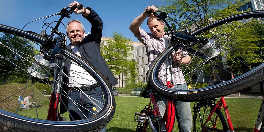 Two men on a city green hold their bicycles on their handlebars.