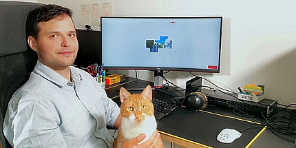 Young man in white shirt with cat on his lap at computer workstation..