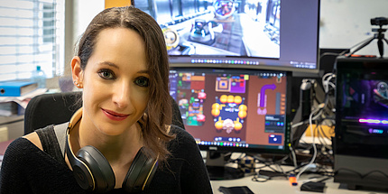 A woman with headphones around her neck sits in front of computer screens running video games.