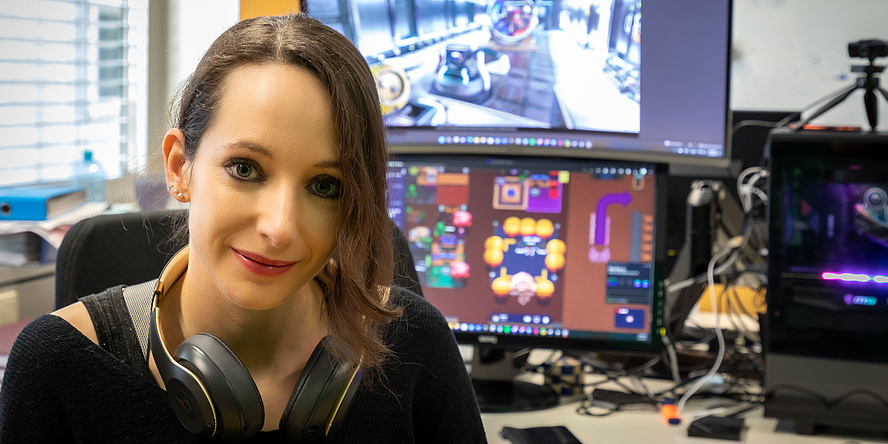 A woman with headphones around her neck sits in front of computer screens running video games.