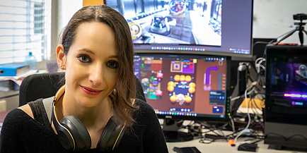 Eine Frau mit Kopfhörern um den Hals sitzt vor Computerbildschirmen, wo Videogames laufen.