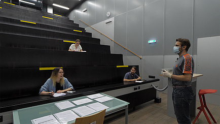 Studierende in einem Hörsaal, Vortragender trägt einen Mund-Nasen-Schutz