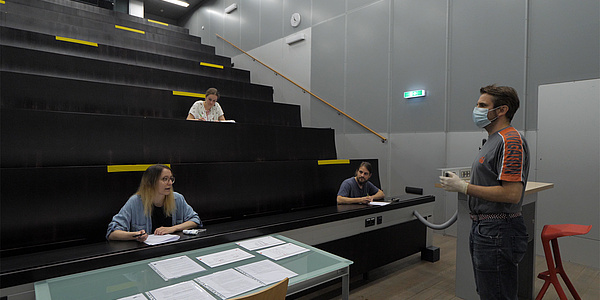 Studierende in einem Hörsaal, Vortragender trägt einen Mund-Nasen-Schutz