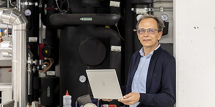 René Rieberer steht in seinem Labor und hält einen Laptop in der Hand. Hinter ihm sind schwarze Rohre zu sehen.