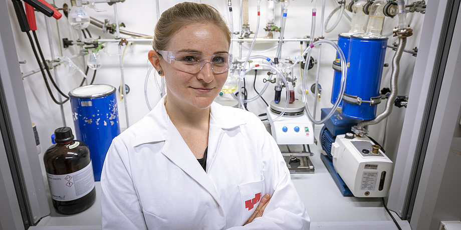 Eine junge Frau mit blonden Haaren steht vor einer Laborbox, in der sich Kabel und Reagenzgläser befinden. Sie trägt einen weißen Labormantel, eine Schutzbrille und hat die Hände verschränkt.