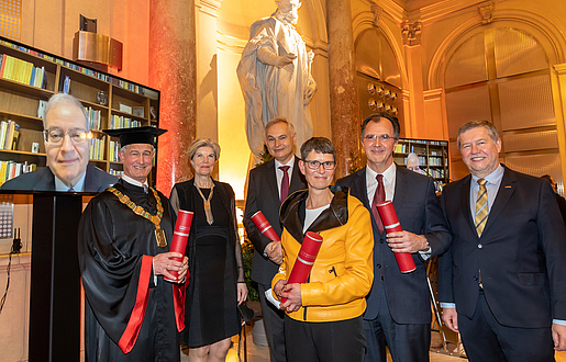 Festively dressed group of people with scrolls of certificates, one person is watching via television