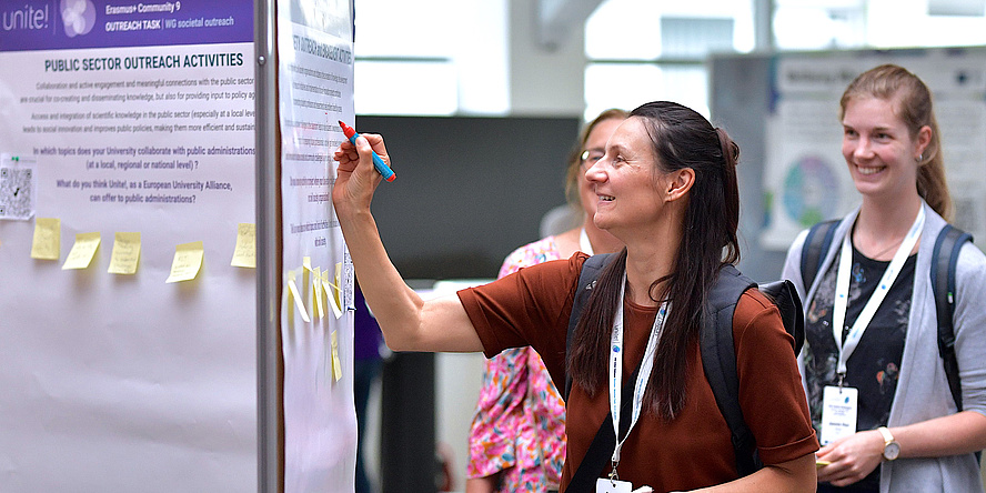 A woman with dark hair and a dark red jumper is writing something on a poster with a pen. Two smiling women can be seen in the background watching the woman write.