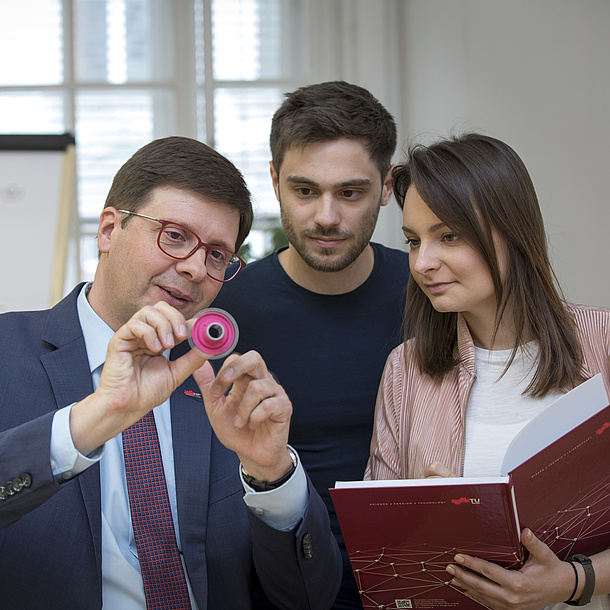 Ein Lehrender erklärt zwei Studierenden ein kleines rundes Objekt.