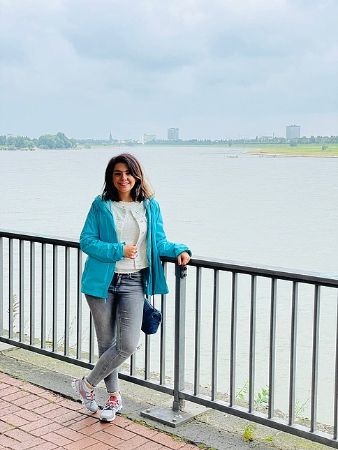 Junge Frau mit blauer Jacke an einer Uferpromenade, städtische Skyline im Hintergrund.