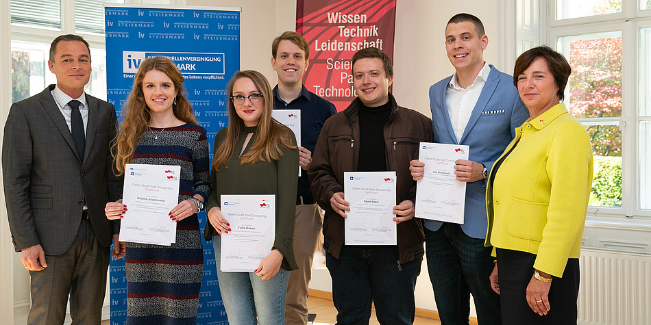 A group of five scholarship holders hold their scholarship certificates in the camera and are flanked by representatives of the sponsors. 
