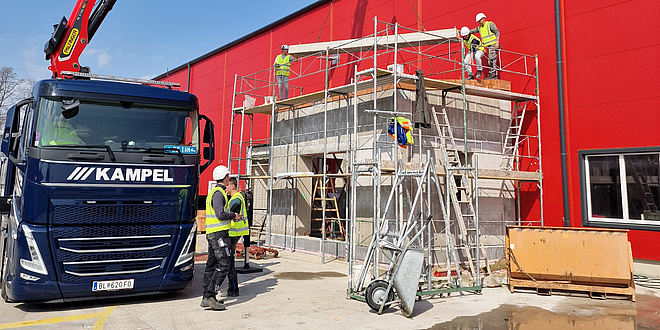 Ein kleines Gebäude ist von einem Gerüst umzäunt. Menschen arbeiten an dem Gebäude. Daneben steht ein Lastwagen mit einem Kran.
