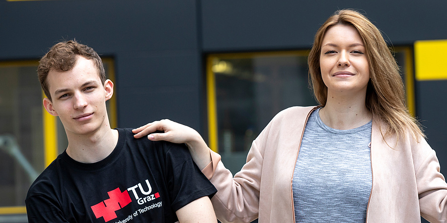 Junger Mann mit schwarzem Shirt mit großem rotem TU Graz-Logo links neben einer jungen Frau mit grauem Shirt und lachsfarbenem Cardigan. Beide blicken direkt in die Kamera, ihre Hand liegt auf seiner Schulter.