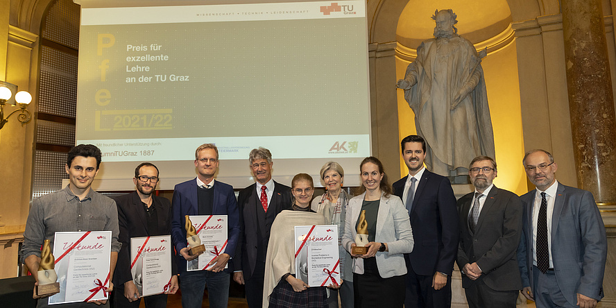 10 lächelnde Menschen stehen vor einer Leinwand, auf die "Preis für exzellente Lehre" projiziert wird. Vier Personen halten eine Siegerurkunde, eine hält eine Trophäe.