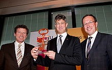Drei Männer in schwarzen Anzügen blicken in die Kamera. Der Mann in der Mitte hält einen großen, orangen Schlüssel in die Kamera. 
