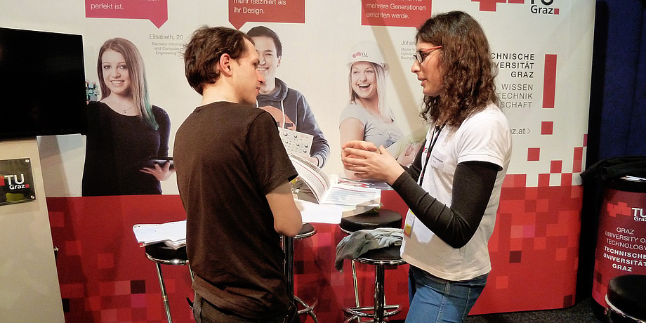 Am TU Graz-Infostand auf der Messe für Beruf, Studium und Weiterbildung in Wien sind eine junge Frau einen Interessent ins Gespräch vertieft.