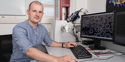 A man with a blue shirt is sitting in front of a screen with a microscope image of a metal powder - many round balls - on it. 