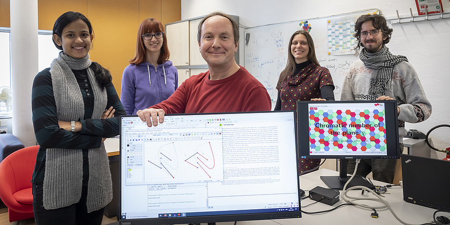 Die Arbeitsgruppe von Oswin Aichholzer steht in einem Büro. Vor ihnen sind zwei Bildschirm mit mathematischen Zeichnungen zu sehen.