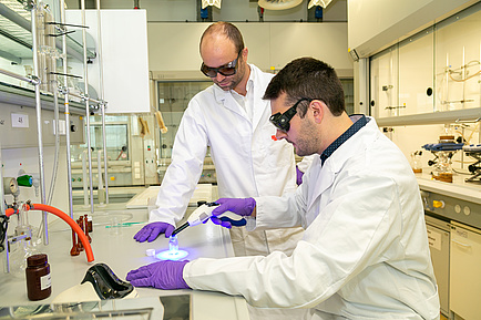 Two men in lab coats and protective goggles, one holding a small light source to a glass object.