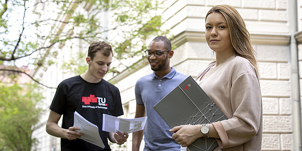 Eine junge Frau blickt in die Kamera, sie hält eine TU Graz-Mappe in der Hand. Im Hintergrund blicken zwei junge Männer auf ein Blatt Papier, einer der beiden trägt ein TU Graz-Shirt.