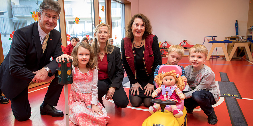 Drei Erwachsene und drei Kinder umgeben von Spielsachen im Turnsaal einer Kinderbetreuungseinrichtung