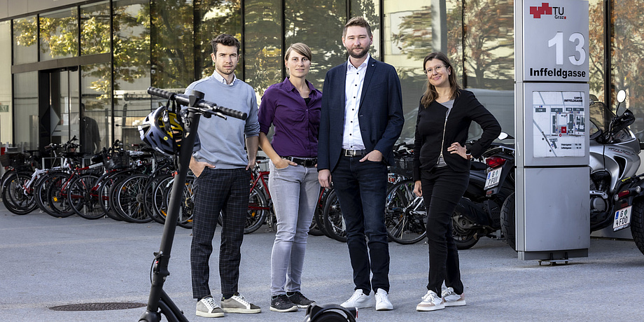 Four people, two women and two men, look into the camera. An e-scooter stands in front of them.