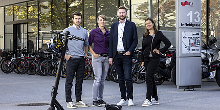 Four people, two women and two men, look into the camera. An e-scooter stands in front of them.