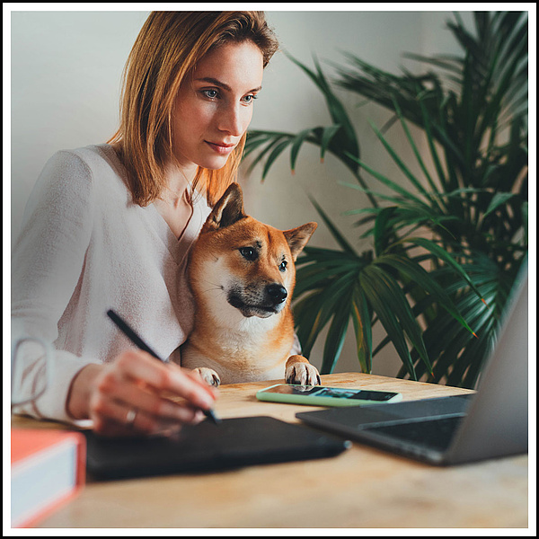 Eine Frau sitzt mit einem Hund an einem Tisch. Vor ihr steht ein aufgeklappter Laptop.