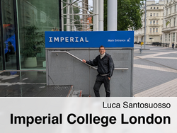 Luca Santosuosso lächelt und steht an einem Treppengeländer vor einem blauen Schild mit der Aufschrift „IMPERIAL Main Entrance“ und dem Logo des Imperial College London. Im Hintergrund sind Gebäude und ein Fahrrad zu sehen.
