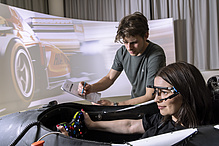 A woman is sitting in a racing simulator, and a man is standing next to the cockpit.