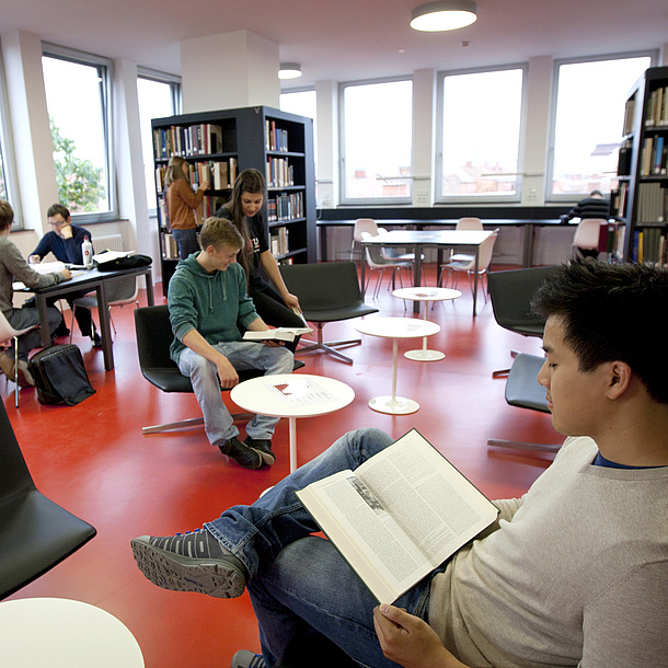 Young people in a room. Some are sitting and reading, others are standing at bookshelves or conversing.