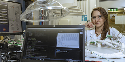 Eine Frau blickt in die Kamera. Sie trägt einen weißen Labormantel und sitzt an einem Tisch. Vor ihr steht ein Laptop, auf dem Diagramme zu sehen sind.