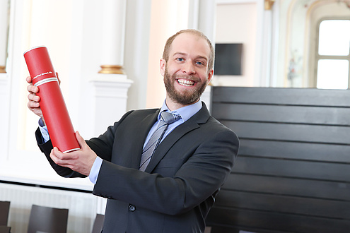 A man holds up a red document roll and smiles into the camera.