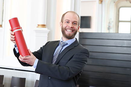 Ein Mann hält eine rote Dokumentenrolle in die Höhe und lächelt in die Kamera
